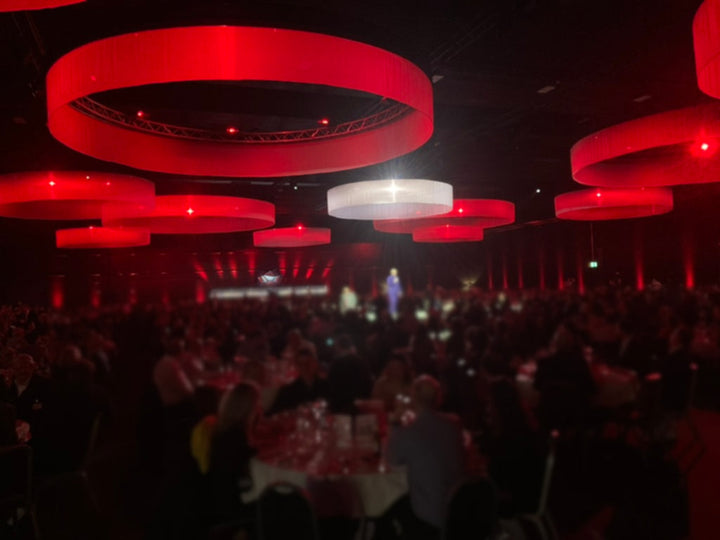 Festsaal mit großen dekorativen rot und weiß ausgeleuchteten Kreisen an der Hallendecke, im unteren Bereich sitzen Personen an Tiaschen und blicken zu einem Moderator in der Mitte des Raumes