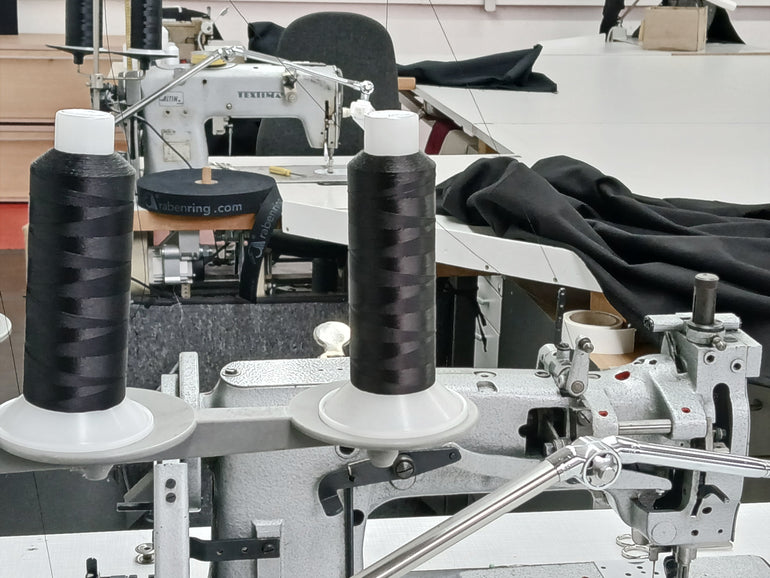 Detailansicht eine Nähmaschine mit schwarzen Garnspulen, im Hintergrund eine weitere Nähmaschine